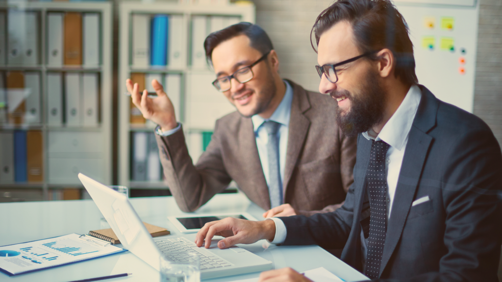 Dois profissionais trabalham sentados à mesa, em frente ao computador, analisando o mercado de logística.