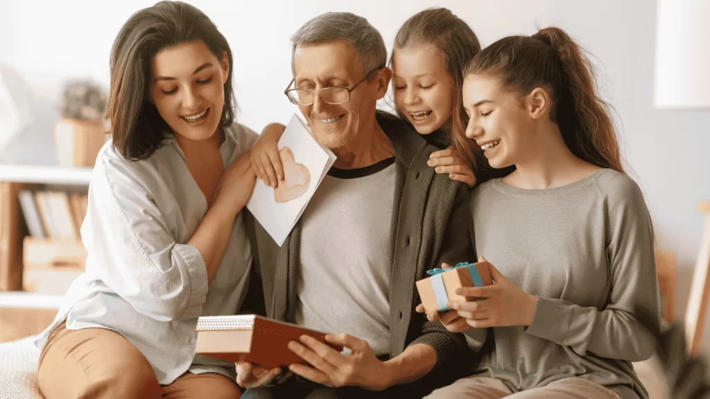 Um homem mais velho, de cabelos grisalhos e óculos, está sentado sorrindo enquanto recebe presentes e carinho de três mulheres mais jovens. À esquerda, uma mulher adulta segura um caderno ou diário; ao fundo, uma menina segura um cartão com um coração desenhado; à direita, uma jovem entrega uma pequena caixa de presente com laço azul. Todos estão sorrindo e demonstram afeto, em um ambiente interno acolhedor e iluminado. A cena transmite uma sensação de celebração familiar, carinho e gratidão.