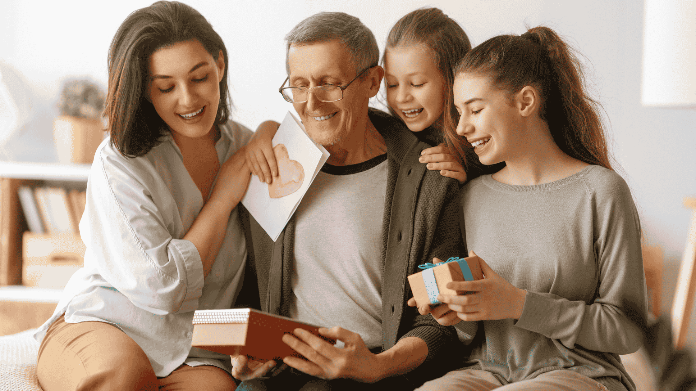 Um homem mais velho, de cabelos grisalhos e óculos, está sentado sorrindo enquanto recebe presentes e carinho de três mulheres mais jovens. À esquerda, uma mulher adulta segura um caderno ou diário; ao fundo, uma menina segura um cartão com um coração desenhado; à direita, uma jovem entrega uma pequena caixa de presente com laço azul. Todos estão sorrindo e demonstram afeto, em um ambiente interno acolhedor e iluminado. A cena transmite uma sensação de celebração familiar, carinho e gratidão.