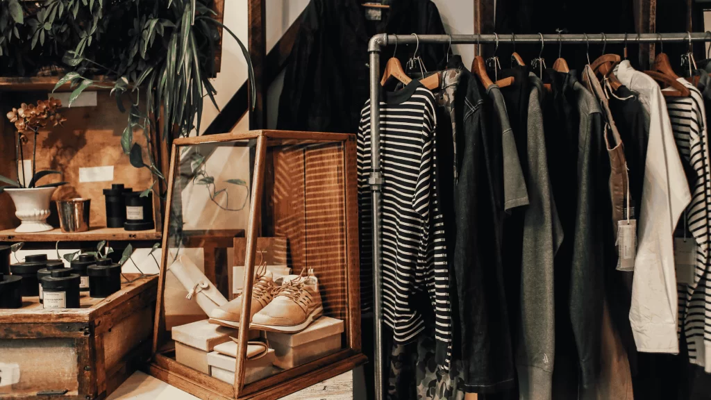  Interior de uma loja ou brechó com atmosfera rústica e aconchegante. À direita, há um varal de roupas metálico com várias peças penduradas em cabides de madeira — a maioria em tons neutros, como preto, branco, cinza e bege, com destaque para listras e tecidos leves. À esquerda, há uma vitrine de madeira com vidro, onde estão expostos um par de tênis claros e algumas caixas organizadoras. Ao fundo, há prateleiras de madeira com vasos de plantas, velas decorativas pretas e outros itens de decoração. O ambiente transmite uma sensação de estilo vintage e curadoria artesanal.