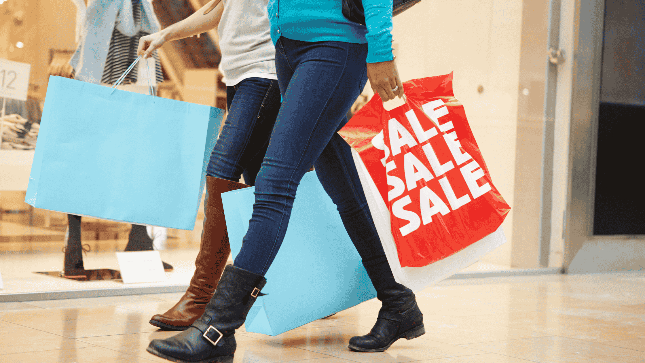 Duas pessoas caminham em um shopping carregando grandes sacolas de compras. Uma delas segura uma sacola vermelha com a palavra "SALE" repetida em branco, indicando liquidação.
