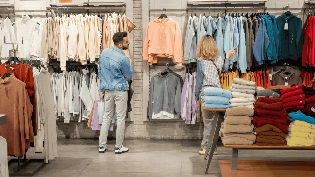 Um homem e uma mulher observam roupas penduradas em uma loja. O ambiente é bem organizado, com peças separadas por cores e tipos, incluindo blusas e moletons, além de mesas com roupas dobradas.