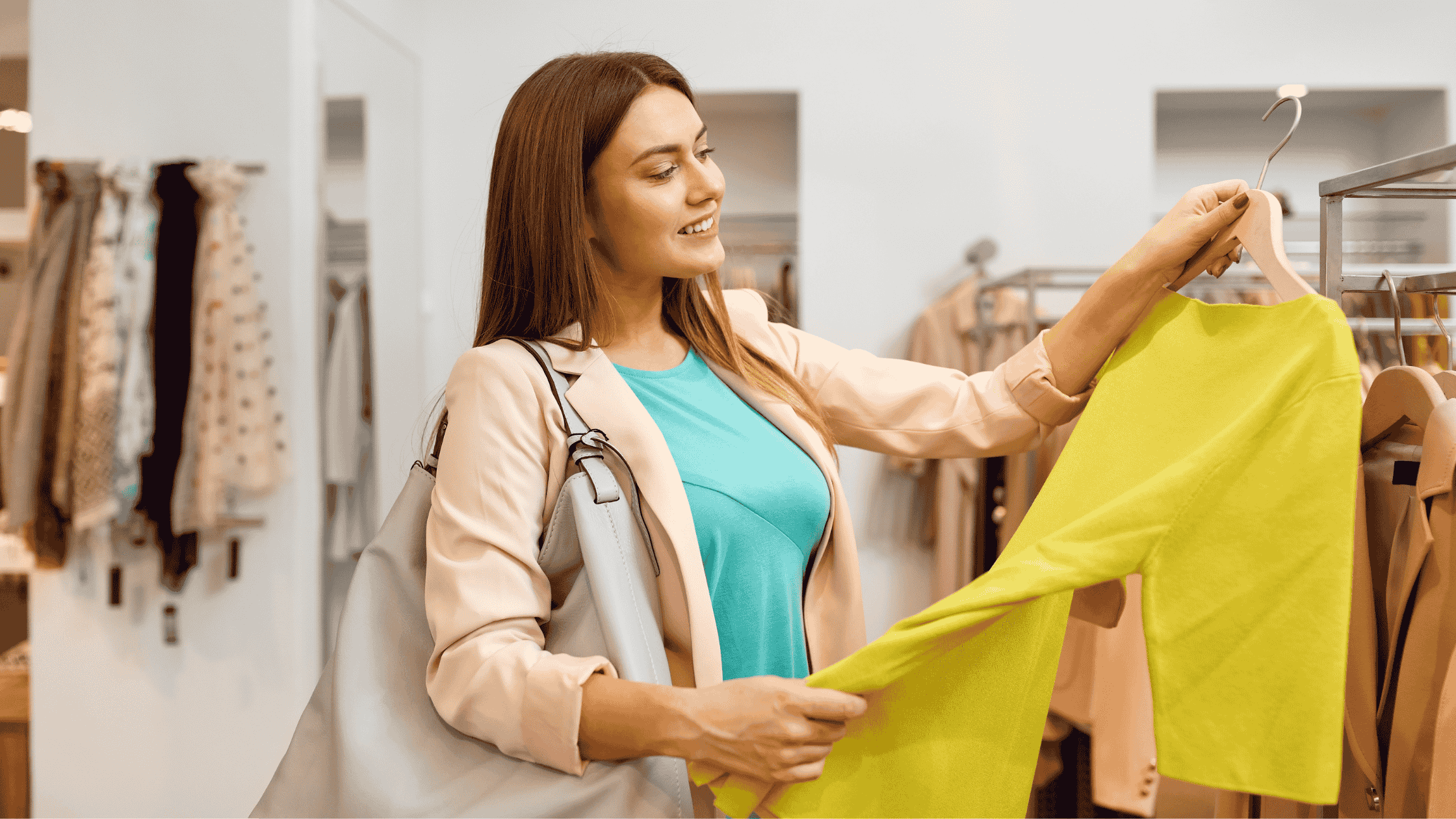 Mulher sorri enquanto observa uma blusa amarela em um cabide, dentro de uma loja de roupas bem iluminada. Ela carrega uma bolsa e parece estar decidindo sobre a compra.