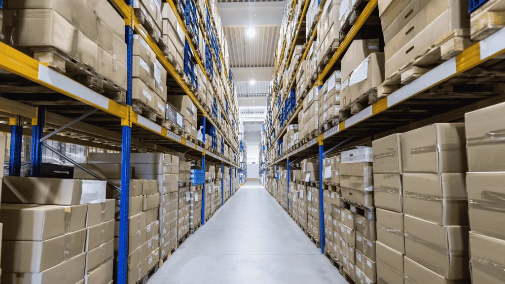 Corredor central de um grande galpão industrial com prateleiras altas repletas de caixas de papelão empilhadas e organizadas, indicando um centro de armazenamento ou distribuição.