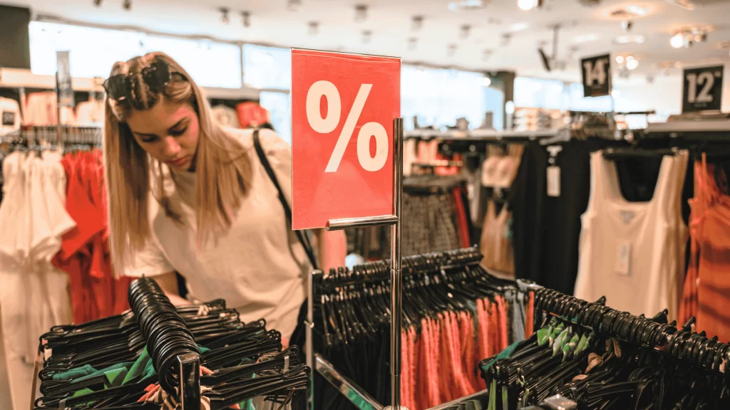 Mulher comprando em uma loja de roupas em promoção, onde aparece uma placa vermelha de desconto