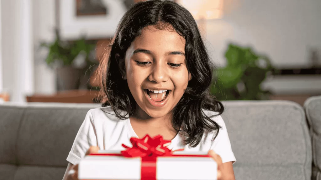 Criança sorrindo de boca aberta para uma caixa de presente que está em suas mãos